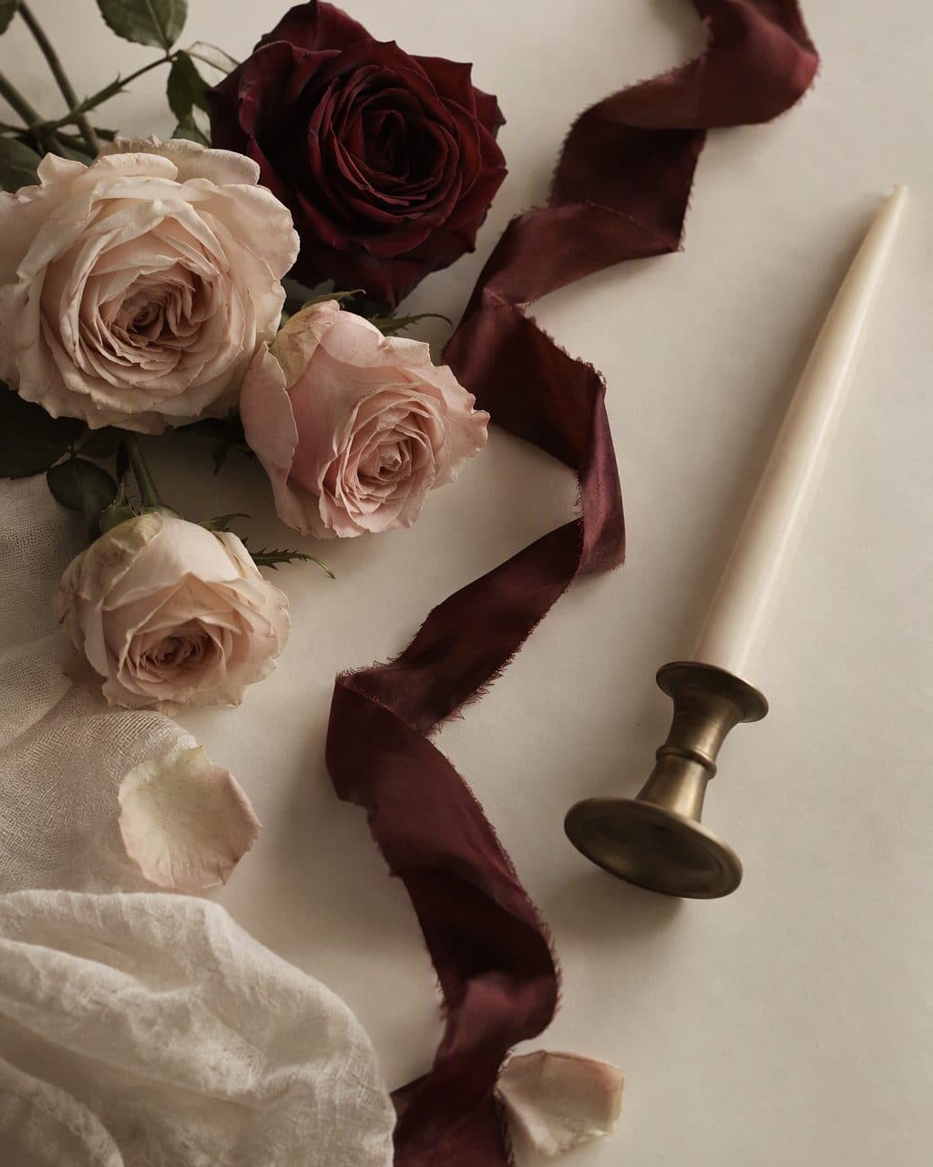Nude roses, burgundy ribbon and a bronze candlestick on ivory paper, seen from above