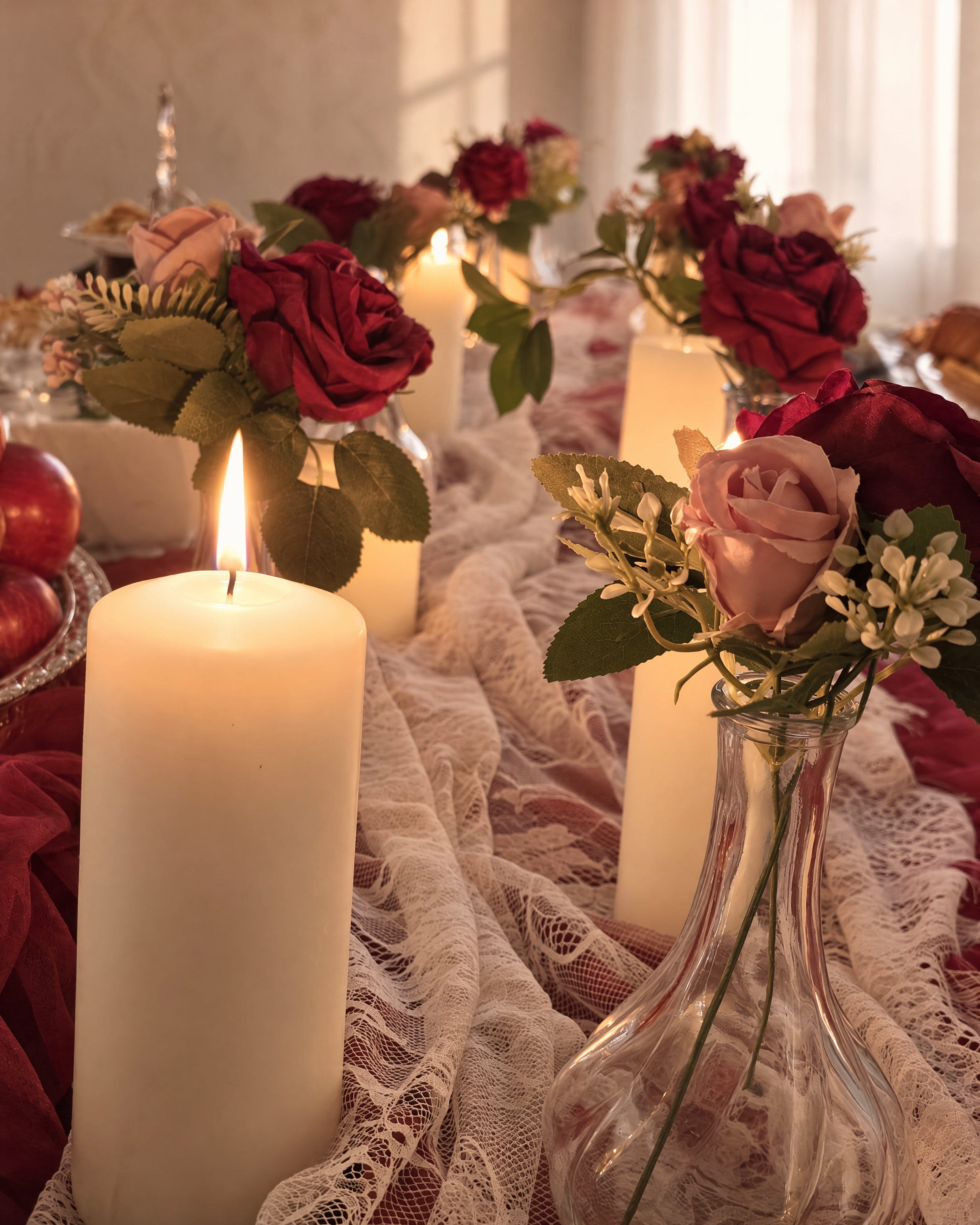 A lit pillar candle close on the lace runner, a blush rose and a burgundy rose in bud vases beside it