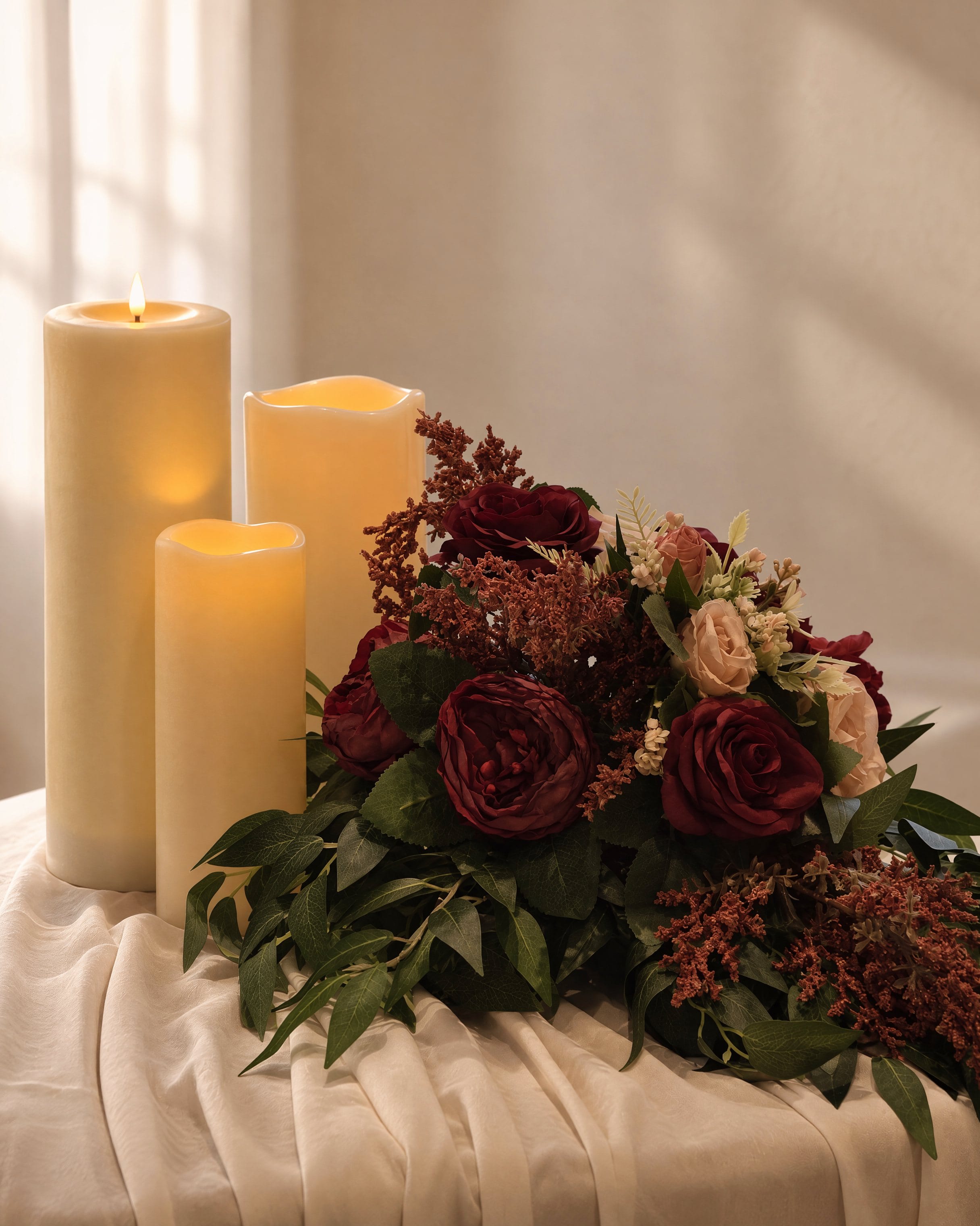 Two ivory pillar candles beside burgundy roses and foliage on a draped console
