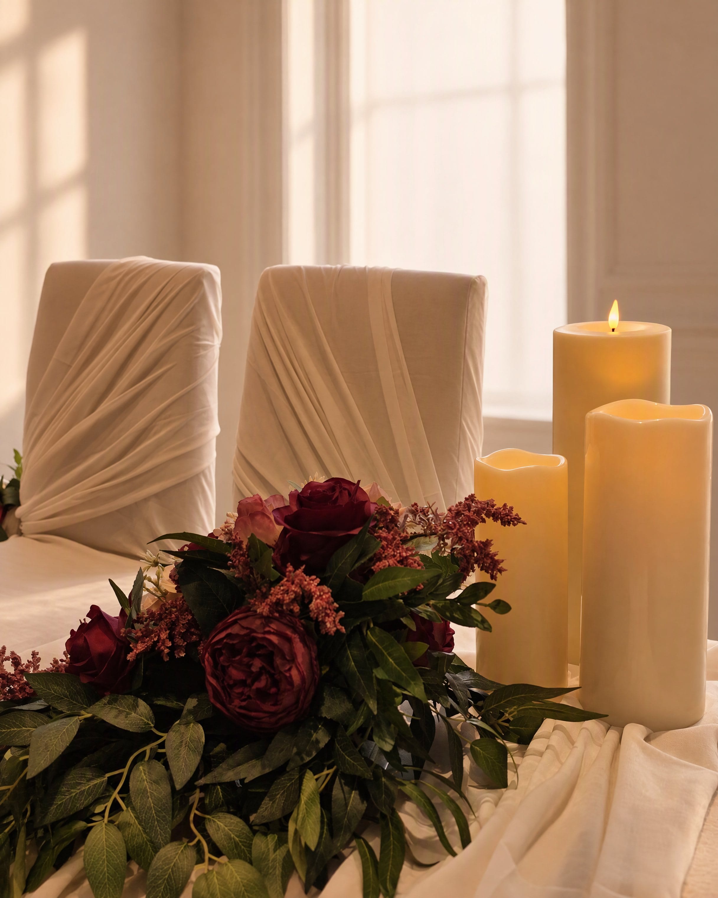 Ivory-draped ceremony chairs behind a low run of burgundy roses and foliage with three pillar candles