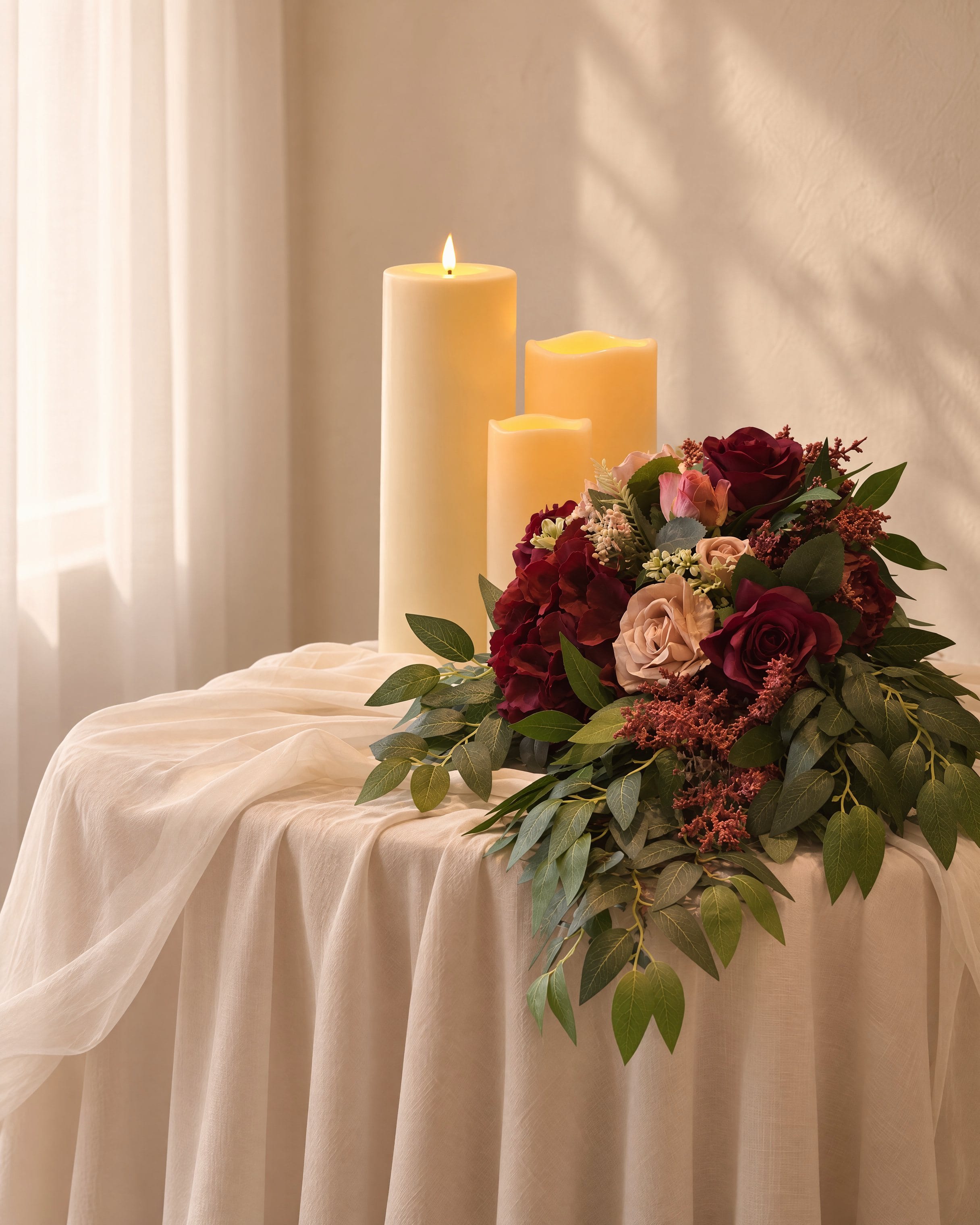 A low arrangement of burgundy roses and foliage with three candles on a draped side table