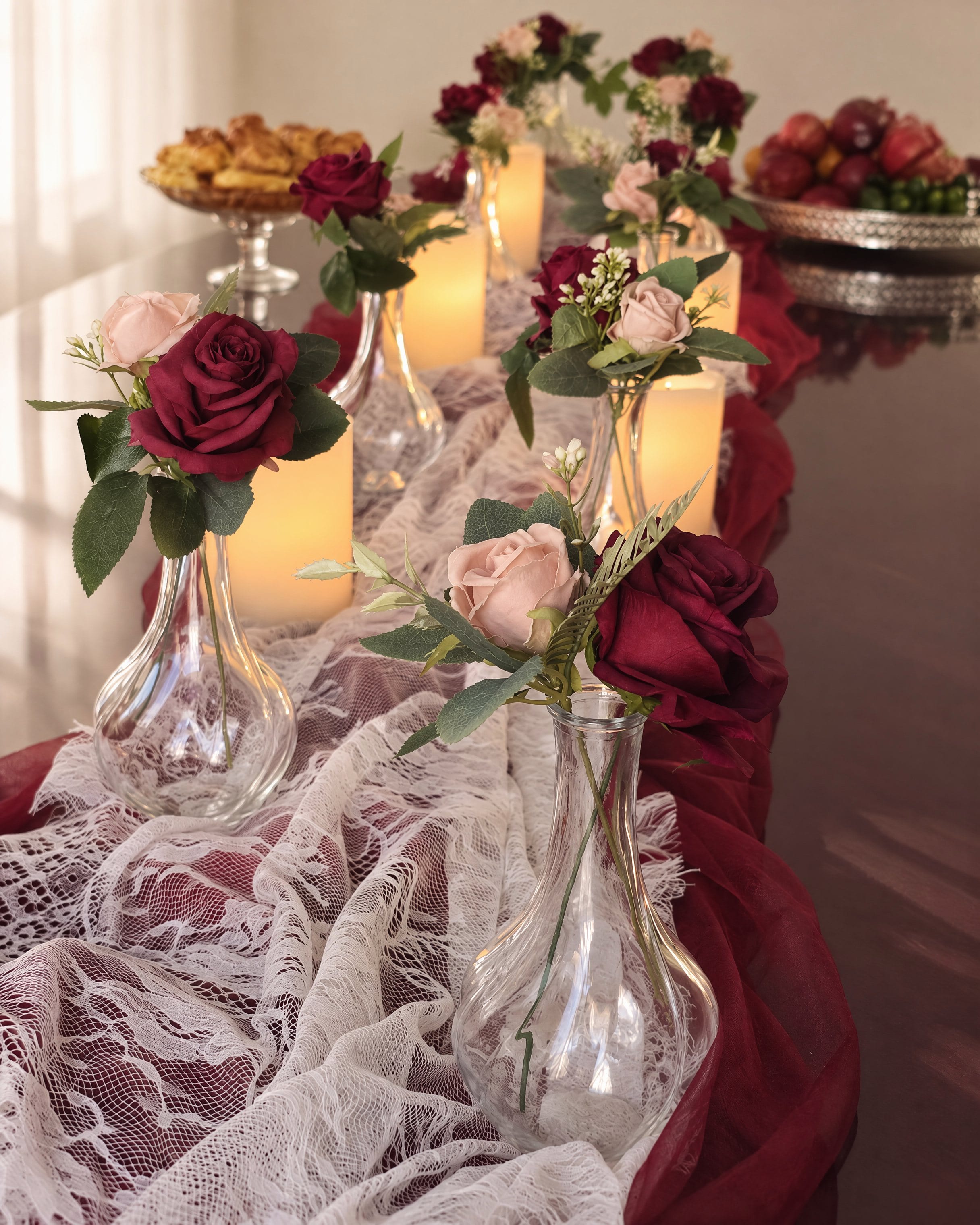 Single burgundy and blush roses in glass bud vases down a lace runner, pillar candles between them